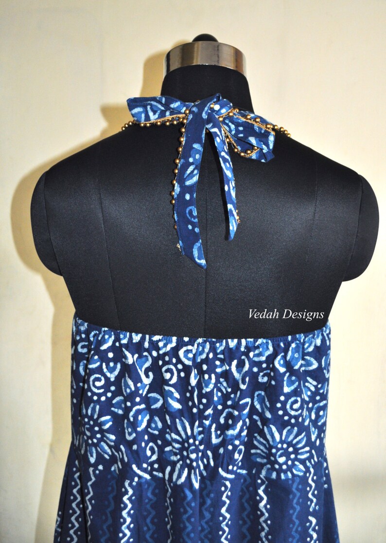 May include: A blue and white patterned halter dress displayed on a black mannequin. The dress features a bow at the neck with gold bead accents. The fabric has a floral and abstract design. The text "Veda Designs" is visible.