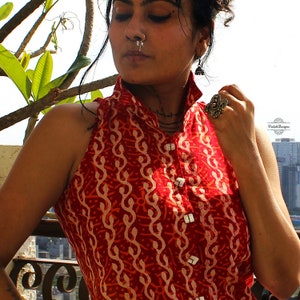 May include: A sleeveless red top featuring a repeating cream-colored wavy pattern and a high collar. The garment has a front button placket with square white buttons. A silver nose ring and a large silver ring are visible on the wearer.