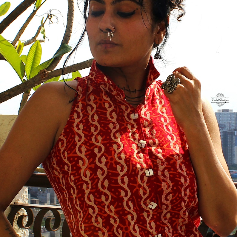 May include: A sleeveless red top featuring a repeating cream-colored wavy pattern and a high collar. The garment has a front button placket with square white buttons. A silver nose ring and a large silver ring are visible on the wearer.