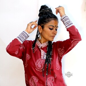 May include: A woman wearing a red long-sleeved shirt with a white paisley print. The shirt has blue and white trim on the cuffs. She is wearing a black beaded necklace and has her hair in a braided bun.