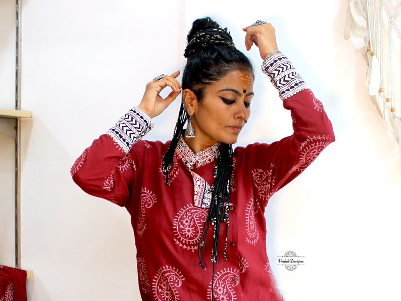 May include: A woman wearing a red long-sleeved shirt with a white paisley print. The shirt has blue and white trim on the cuffs. She is wearing a black beaded necklace and has her hair in a braided bun.