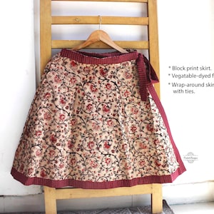 Op de afbeelding: Een wikkelrok van beige stof met een rood en zwart bloemenpatroon in blokdruk. De rok heeft een effen donkerrode tailleband en zoom, en is voorzien van strikbanden. De tekst op de afbeelding luidt: Block print skirt, Vegetable-dyed fabric, Wrap-around skirt with ties.