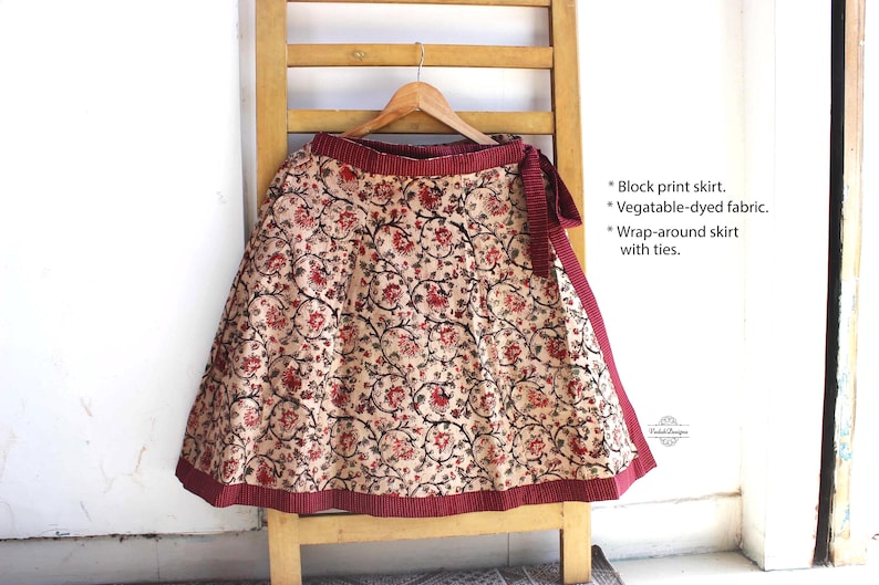 Op de afbeelding: Een wikkelrok van beige stof met een rood en zwart bloemenpatroon in blokdruk. De rok heeft een effen donkerrode tailleband en zoom, en is voorzien van strikbanden. De tekst op de afbeelding luidt: Block print skirt, Vegetable-dyed fabric, Wrap-around skirt with ties.