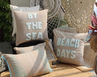 May include: Four decorative throw pillows with embroidered text. The pillows are beige with light blue embroidery. The text on the pillows reads "By the Sea", "Beach Days", and two pillows with a leaf pattern.