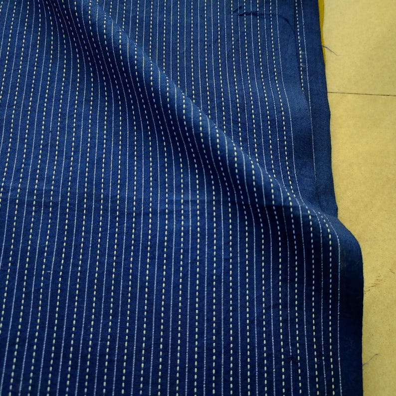 May include: A deep blue fabric featuring a repeating pattern of vertical white dashed lines. The textile has a visible weave texture and is draped to show folds, highlighting the consistent alignment of the white dashes across the dark surface.