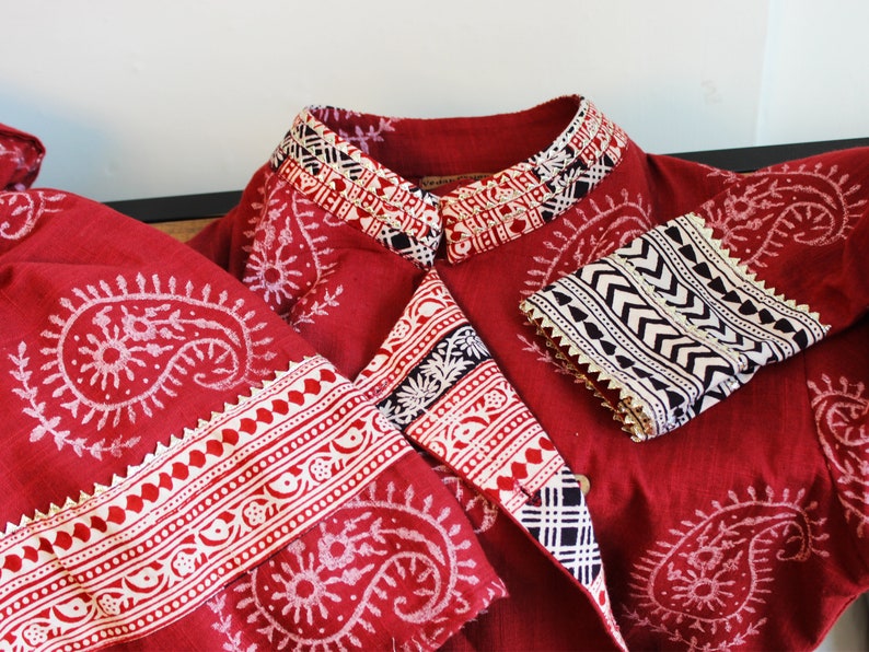 May include: A red cotton jacket with a white and black paisley print. The jacket has a mandarin collar and long sleeves with white and black trim. The jacket is made in India.