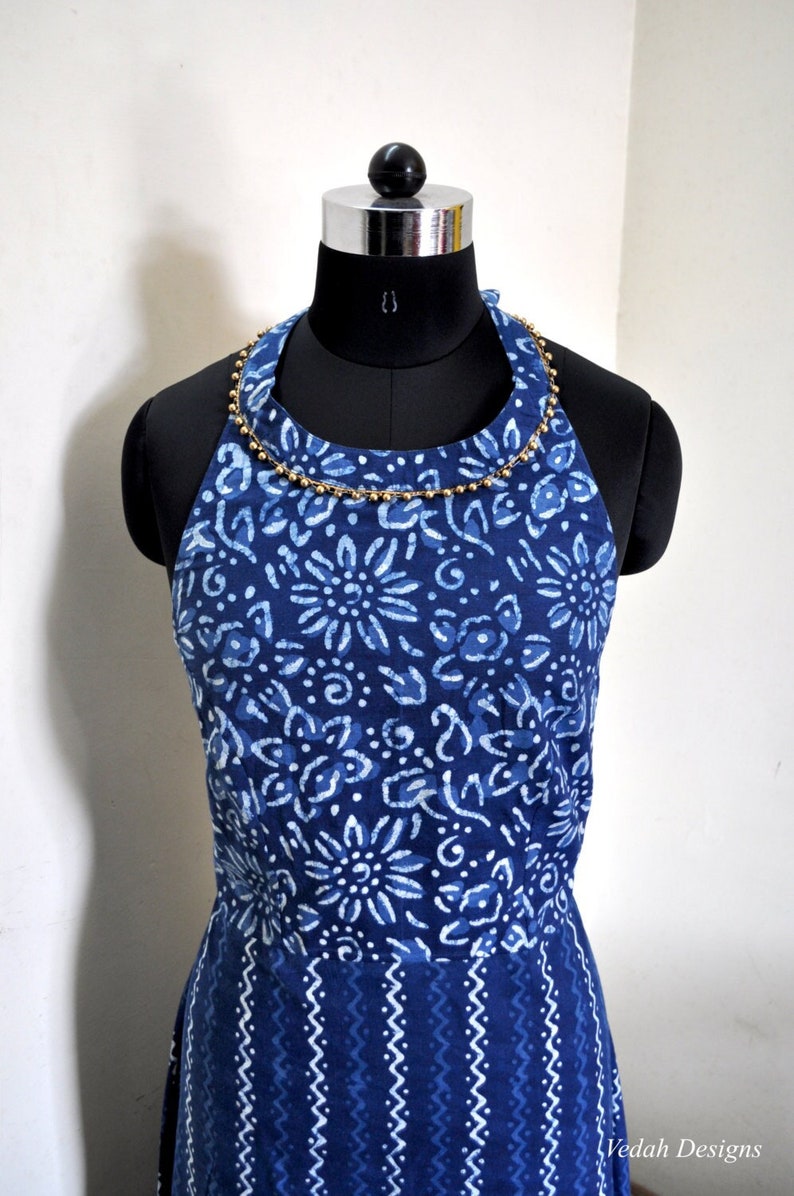 May include: A halter-style dress in indigo blue, adorned with a white floral pattern. The neckline is embellished with a gold beaded trim. The dress is displayed on a black mannequin.