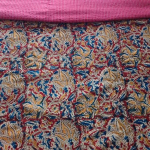 Op de afbeelding: Een textielstof met een dicht bloemenpatroon in tinten rood, geel en blauw op een cr&egrave;mekleurige achtergrond. Het bovenste gedeelte van de stof is effen magenta met dunne, horizontale witte stiklijnen die over de breedte van de stof lopen.