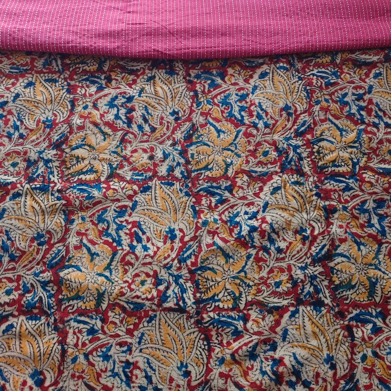 Op de afbeelding: Een textielstof met een dicht bloemenpatroon in tinten rood, geel en blauw op een cr&egrave;mekleurige achtergrond. Het bovenste gedeelte van de stof is effen magenta met dunne, horizontale witte stiklijnen die over de breedte van de stof lopen.