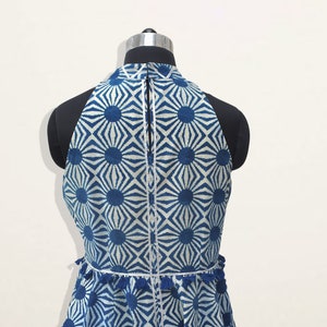 May include: A sleeveless top featuring a blue and white geometric sunburst pattern. The garment has a high neckline, a vertical center zipper, and a horizontal waist seam accented with small blue tassels and a thin silver metallic trim.