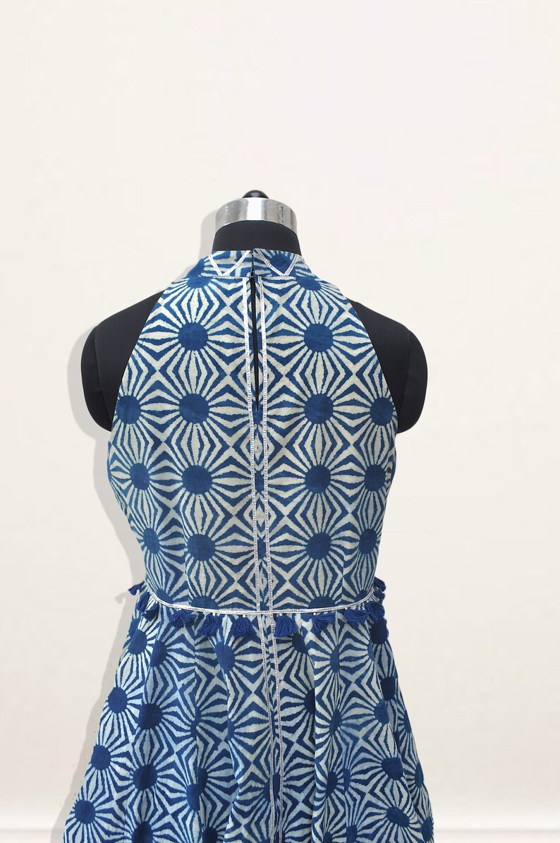 May include: A sleeveless top featuring a blue and white geometric sunburst pattern. The garment has a high neckline, a vertical center zipper, and a horizontal waist seam accented with small blue tassels and a thin silver metallic trim.
