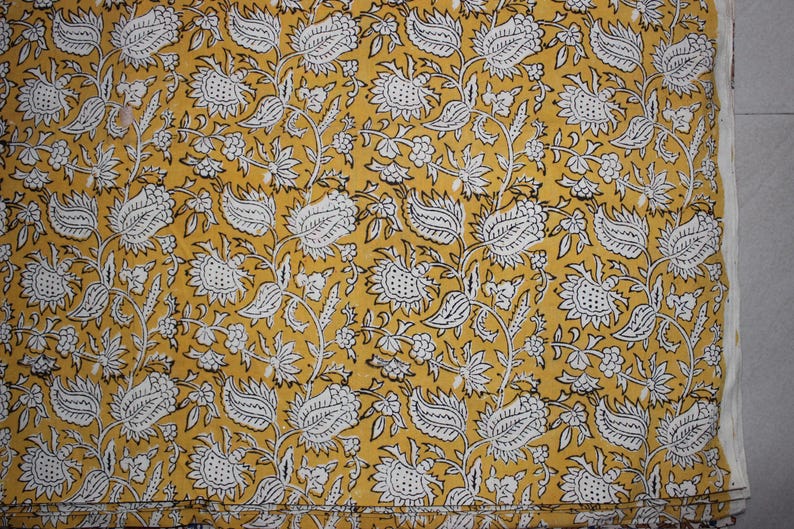 May include: A mustard yellow fabric featuring a repeating pattern of white floral and leaf motifs outlined in thin black lines. The design consists of stylized blossoms and foliage arranged in a dense, continuous botanical print across the surface.