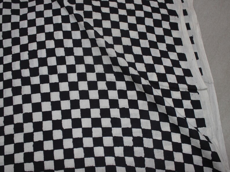 May include: A piece of fabric featuring a repeating black and white checkered pattern. The design consists of uniform square shapes arranged in a grid, with the black squares showing slight variations in their edges and texture across the surface.