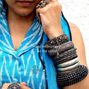 K&ouml;nnte beinhalten: Eine Frau tr&auml;gt ein blau-wei&szlig; gemustertes Hemd mit silbernen Armreifen am Handgelenk. Das Hemd hat wei&szlig;e Stickereien am Kragen. Der Text "Hand embroidery on the collar" ist auf dem Bild zu sehen. Der Text "Vedak Designs" ist ebenfalls auf dem Bild zu sehen.
