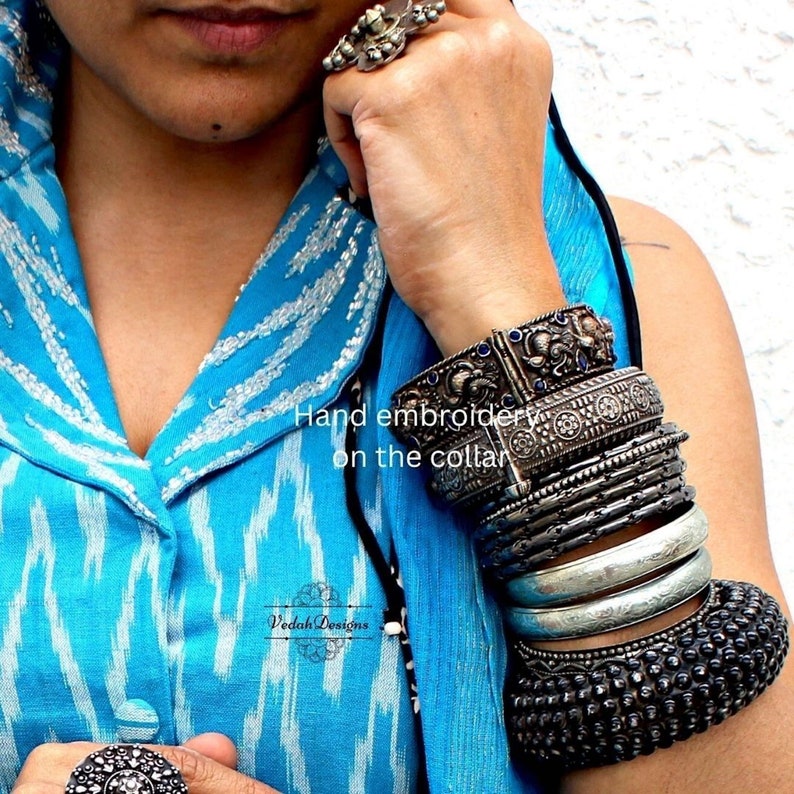 K&ouml;nnte beinhalten: Eine Frau tr&auml;gt ein blau-wei&szlig; gemustertes Hemd mit silbernen Armreifen am Handgelenk. Das Hemd hat wei&szlig;e Stickereien am Kragen. Der Text "Hand embroidery on the collar" ist auf dem Bild zu sehen. Der Text "Vedak Designs" ist ebenfalls auf dem Bild zu sehen.