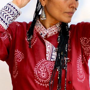 May include: A red cotton shirt with a white paisley print and a white collar. The shirt has long sleeves with white and blue trim. The model is wearing the shirt with black braids.