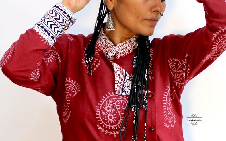 May include: A red cotton shirt with a white paisley print and a white collar. The shirt has long sleeves with white and blue trim. The model is wearing the shirt with black braids.