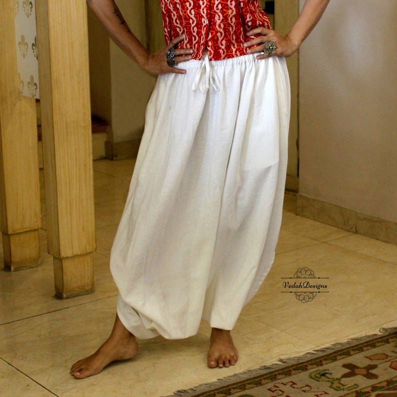 May include: Solid white harem pants featuring a gathered elastic waistband with a functional drawstring tie. The pants have a loose, voluminous silhouette that tapers toward the ankles. A small logo with the text VedahDesigns is visible.