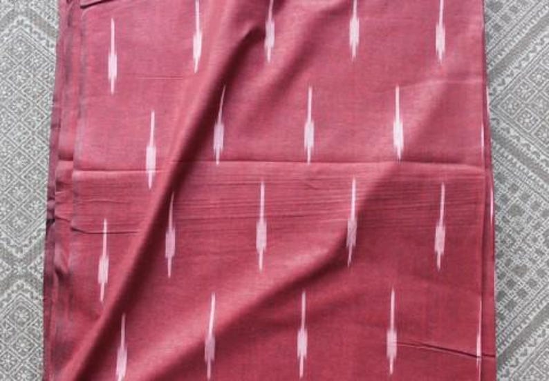 May include: A deep maroon fabric featuring a repeating pattern of vertical, light pink ikat-style dashes. The textile has a visible woven texture with subtle horizontal lines running across the surface, creating a consistent and structured appearance.