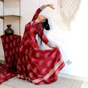 May include: A woman in a maroon paisley print dress with white trim is performing a dance pose. The dress has a long skirt and a fitted bodice. The woman is wearing her hair in a braid and has a natural makeup look.