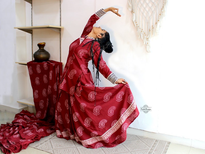 May include: A woman in a maroon paisley print dress with white trim is performing a dance pose. The dress has a long skirt and a fitted bodice. The woman is wearing her hair in a braid and has a natural makeup look.