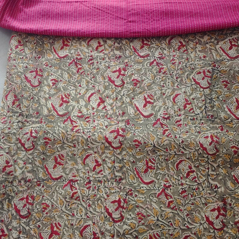 Op de afbeelding: Een textielstuk met een bovenste gedeelte van effen magentakleurige stof met dunne witte horizontale strepen, gelaagd over een groter gedeelte van stof met een dicht, herhalend bloemenpatroon in tinten olijfgroen, rood en geel.