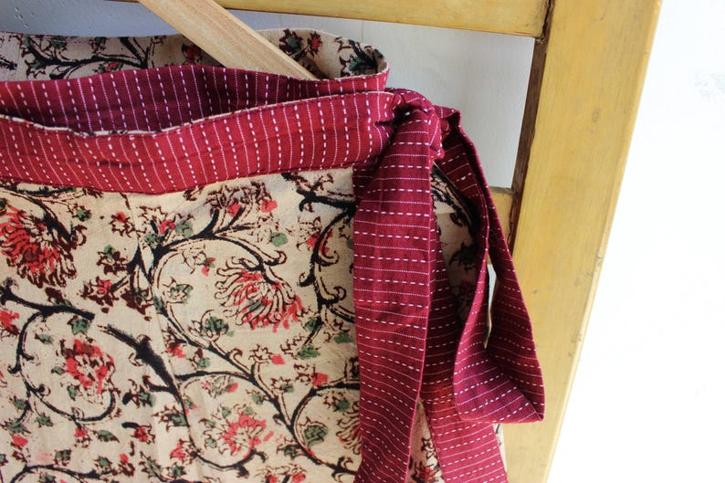 Op de afbeelding: Een wikkelrok met een cr&egrave;mekleurige basis en een zwart en rood bloemenrankpatroon. De tailleband en het strikkoord zijn gemaakt van effen donkerrode stof met dunne, horizontale witte stiksels, wat een contrast vormt met de bloemenprint.
