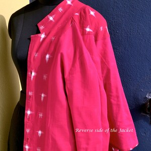 May include: A bright pink jacket featuring a white cross-hatch pattern along the front placket and shoulder. The garment has a button-down front and long sleeves. The text "Reverse side of the Jacket" and "Vedah Designs" is visible on the image.