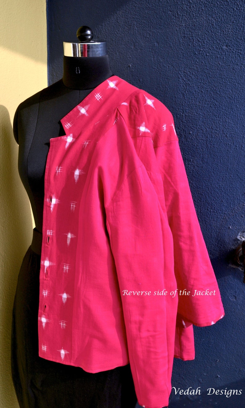 May include: A bright pink jacket featuring a white cross-hatch pattern along the front placket and shoulder. The garment has a button-down front and long sleeves. The text "Reverse side of the Jacket" and "Vedah Designs" is visible on the image.