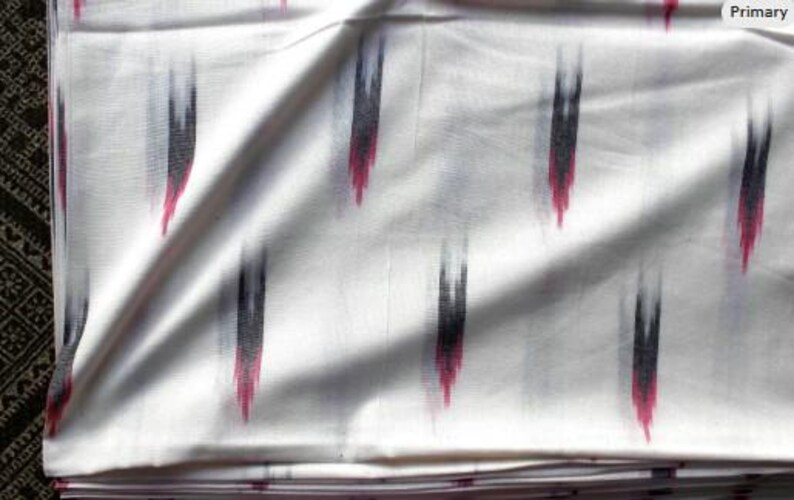 May include: A white fabric featuring a repeating ikat pattern of vertical, blurred brushstroke-like shapes in black and magenta. The textile has a smooth, woven appearance with soft folds that create subtle shadows across the surface of the material.