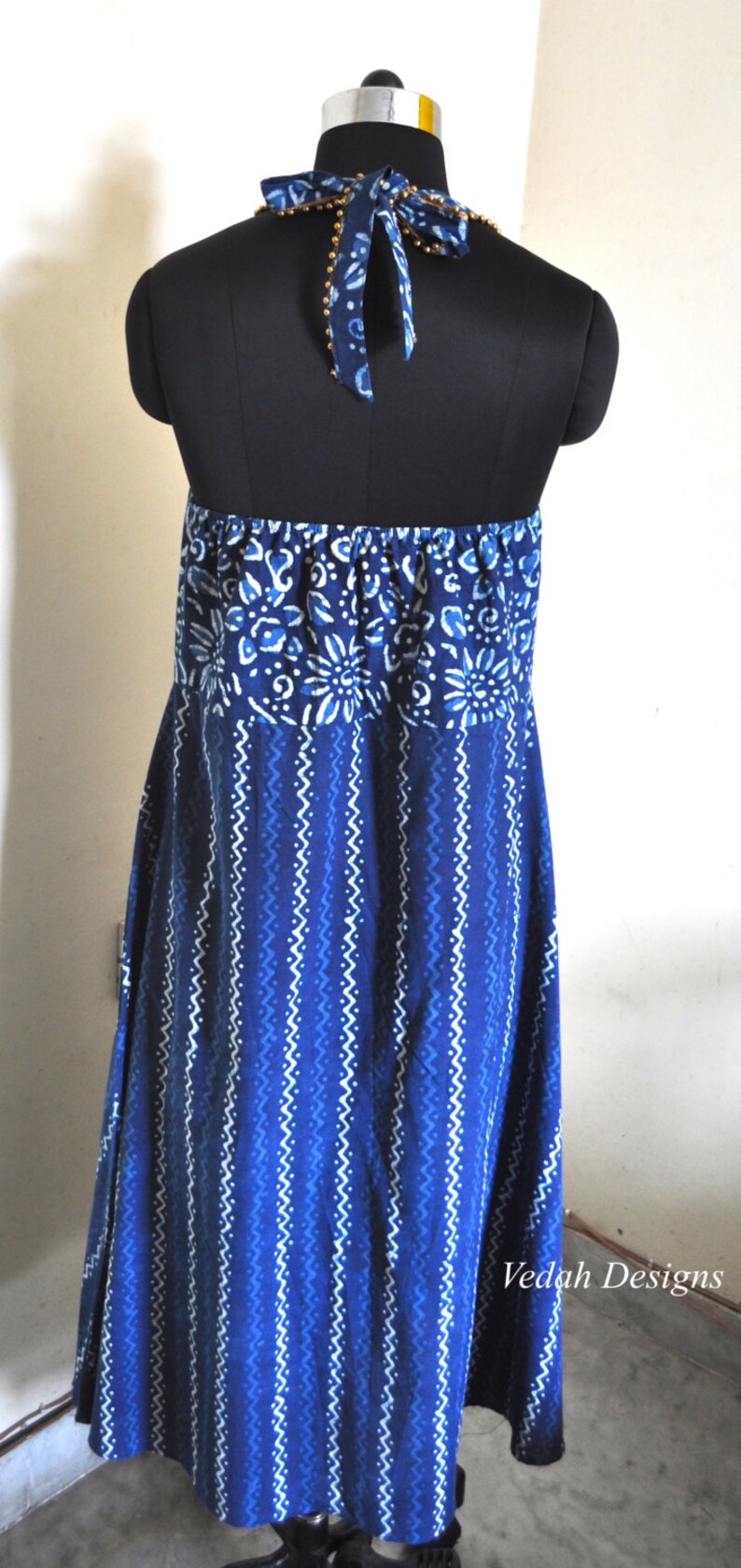 May include: A blue and white patterned halter dress on a black mannequin. The dress features a floral design at the top and a zig-zag pattern on the skirt. The halter tie is adorned with small gold beads.