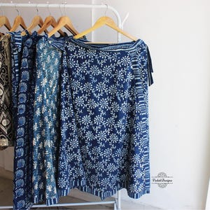 May include: A collection of wrap skirts hanging on wooden hangers on a clothing rack. The skirts feature various blue and white block-printed floral and geometric patterns. A small logo with the text Vedah Designs is visible in the bottom right.