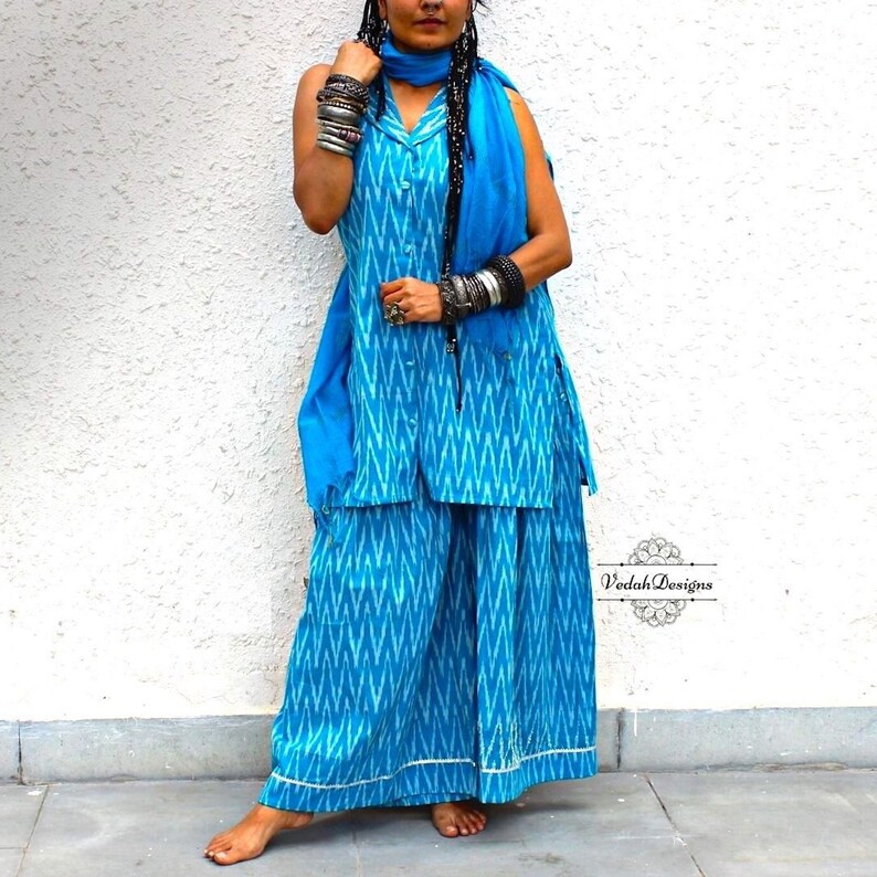 K&ouml;nnte beinhalten: Ein blaues und wei&szlig;es Ikat-Druck-Oberteil ohne &Auml;rmel und eine Palazzo-Hose mit einem passenden blauen Schal. Das Outfit verf&uuml;gt &uuml;ber einen Knopfverschluss und einen wei&szlig;en Saum. "Voodah Designs" ist in der unteren rechten Ecke des Bildes gedruckt.