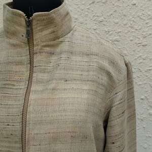 May include: A beige jacket with a textured, slubbed fabric finish featuring subtle horizontal dark gray streaks. The garment includes a high collar, long sleeves, and a central front zipper closure with a matching beige zipper tape and metal pull.
