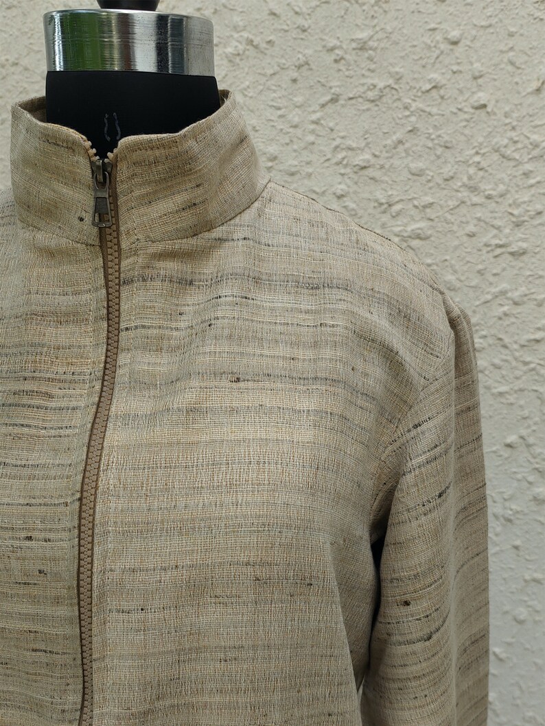 May include: A beige jacket with a textured, slubbed fabric finish featuring subtle horizontal dark gray streaks. The garment includes a high collar, long sleeves, and a central front zipper closure with a matching beige zipper tape and metal pull.