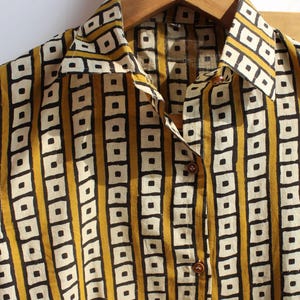 May include: A sleeveless button-down shirt featuring a repeating geometric pattern of black and white squares arranged in vertical stripes against a mustard yellow background, complete with a pointed collar and brown buttons down the front placket.