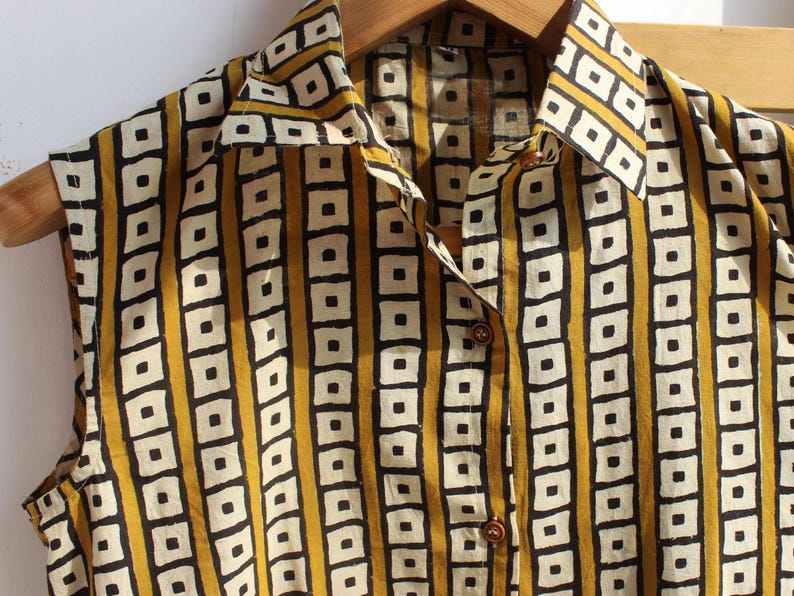 May include: A sleeveless button-down shirt featuring a repeating geometric pattern of black and white squares arranged in vertical stripes against a mustard yellow background, complete with a pointed collar and brown buttons down the front placket.