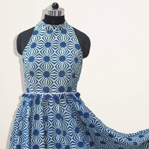May include: A sleeveless dress featuring a high neckline and a blue and white geometric sunburst pattern. The waist is accented with a thin white trim and small blue tassels. The skirt has a flared silhouette that extends to the right side.