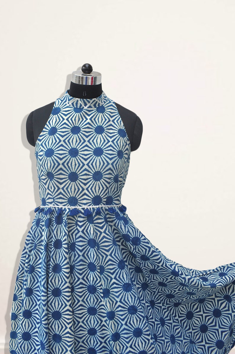 May include: A sleeveless dress featuring a high neckline and a blue and white geometric sunburst pattern. The waist is accented with a thin white trim and small blue tassels. The skirt has a flared silhouette that extends to the right side.