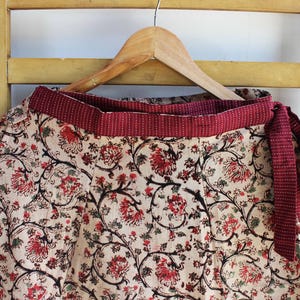Op de afbeelding: Een wikkelrok met een beige basis en een herhalend bloemenpatroon in rood en zwart. De rok heeft een brede, effen donkerrode tailleband met een subtiele witte stippeltextuur en een bijpassende striksluiting aan de zijkant, hangend aan een hanger.