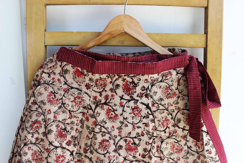 Op de afbeelding: Een wikkelrok met een beige basis en een herhalend bloemenpatroon in rood en zwart. De rok heeft een brede, effen donkerrode tailleband met een subtiele witte stippeltextuur en een bijpassende striksluiting aan de zijkant, hangend aan een hanger.