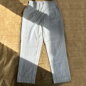 May include: A pair of white trousers featuring an elasticized waistband with a drawstring closure, two side pockets, and a straight-leg silhouette. The fabric has a visible woven texture, and the garment is presented in a flat-lay orientation.