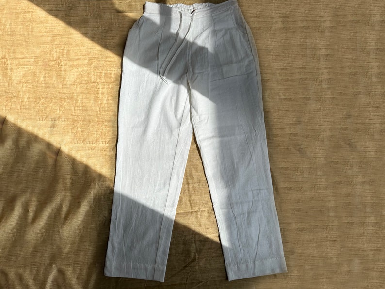May include: A pair of white trousers featuring an elasticized waistband with a drawstring closure, two side pockets, and a straight-leg silhouette. The fabric has a visible woven texture, and the garment is presented in a flat-lay orientation.