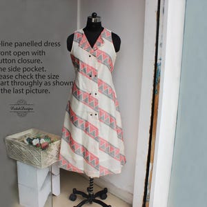 May include: An A-line panelled dress with a front button closure and one side pocket. The dress features a white base with a repeating pattern of pink and gray geometric shapes. The text on the left side of the image reads: "A-line panelled dress, Front open with button closure, One side pocket."