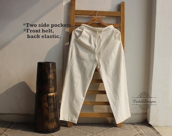 May include: Cream-coloured linen trousers with a drawstring waist, displayed on a wooden hanger. The trousers feature two side pockets and a front belt with back elastic. A tall, dark wooden vase is on the left. Text on the wall reads: "Two side pockets. Front belt, back elastic."
