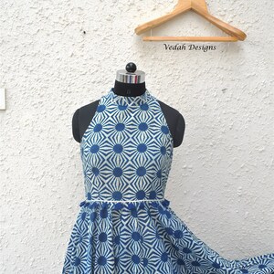May include: A sleeveless dress featuring a high neckline and a blue and white geometric sunburst pattern. The waist is accented with a thin white trim and a row of small blue tassels. The dress is displayed on a mannequin with a wooden hanger.