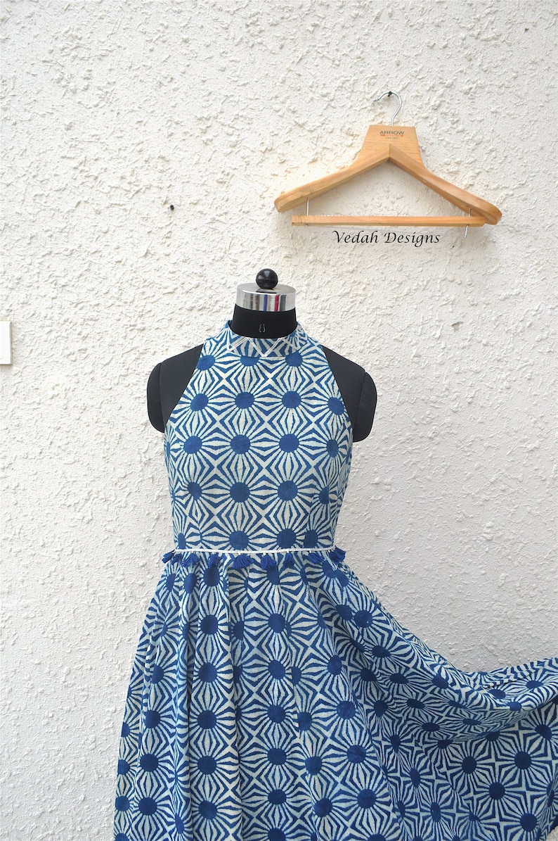May include: A sleeveless dress featuring a high neckline and a blue and white geometric sunburst pattern. The waist is accented with a thin white trim and a row of small blue tassels. The dress is displayed on a mannequin with a wooden hanger.