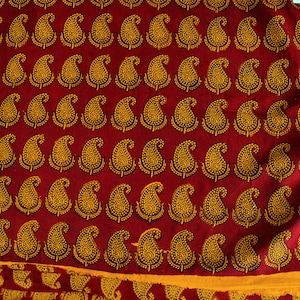 Può includere: Tessuto rosso con un motivo ripetuto di disegni paisley dorati. Il tessuto ha un bordo giallo con un motivo geometrico bianco e nero.
