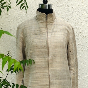 May include: A beige jacket with a textured, horizontal slub pattern, featuring a high collar and a full-length front zipper. The garment has long sleeves and a straight hem, constructed from a woven fabric with subtle variations in light brown tones.