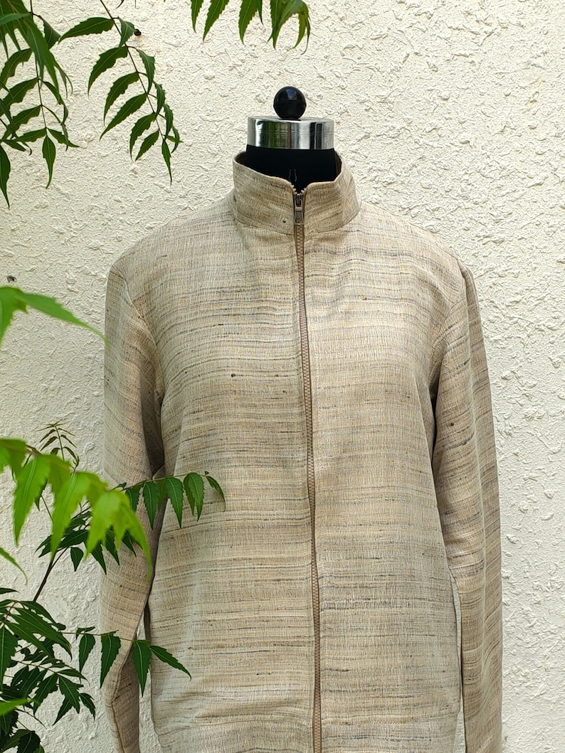 May include: A beige jacket with a textured, horizontal slub pattern, featuring a high collar and a full-length front zipper. The garment has long sleeves and a straight hem, constructed from a woven fabric with subtle variations in light brown tones.